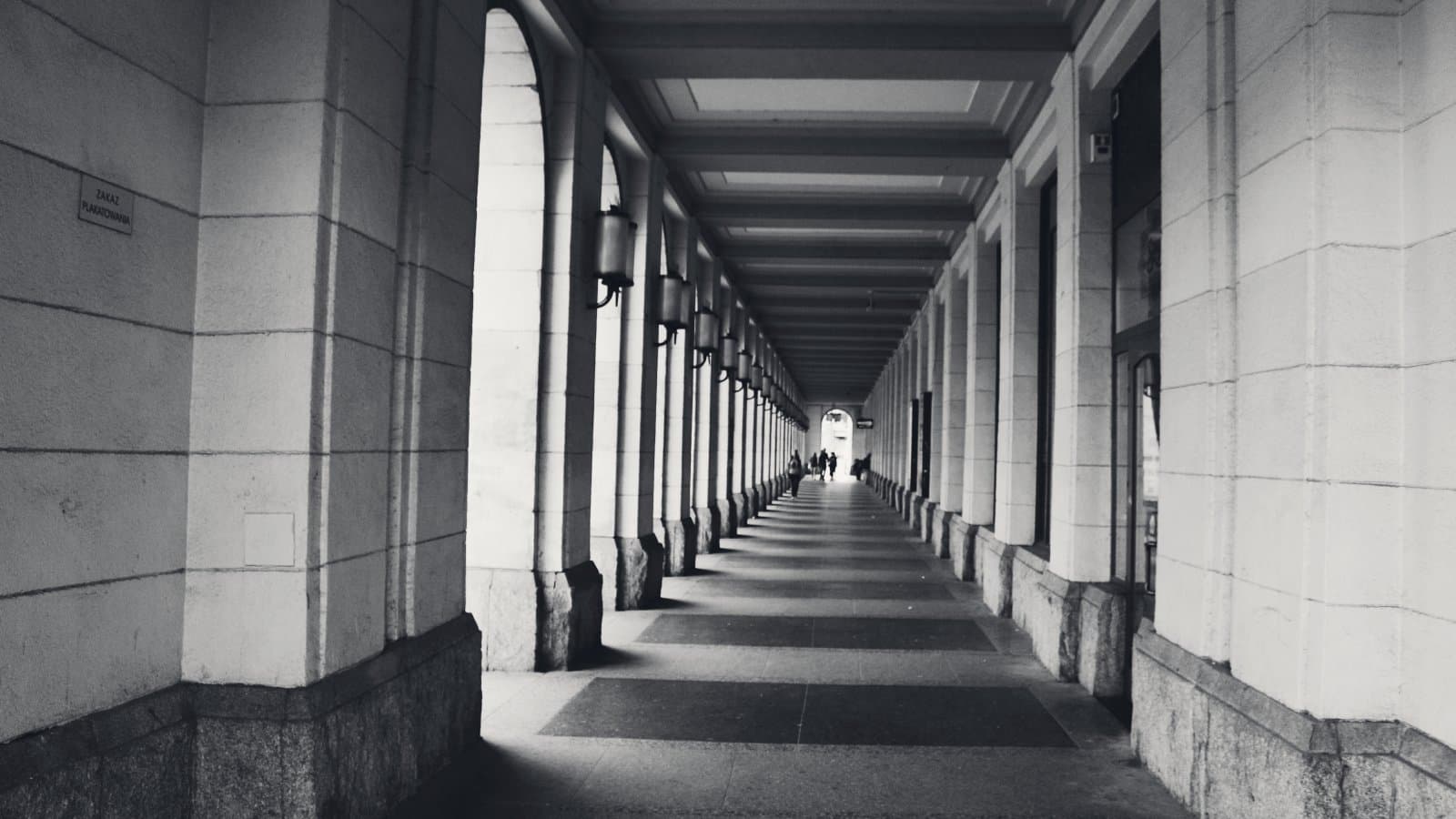 A stone colonnade of repeating arches and columns receding to a distant lit opening, hard side light throwing the column shadows across the paving.