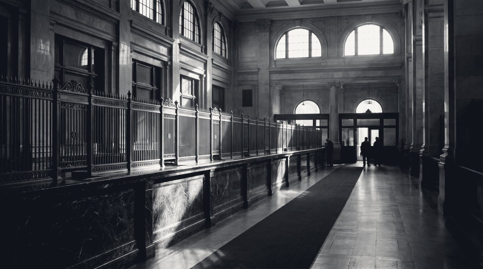 The long marble counter run and teller screens of a grand early-century banking hall in raking light.
