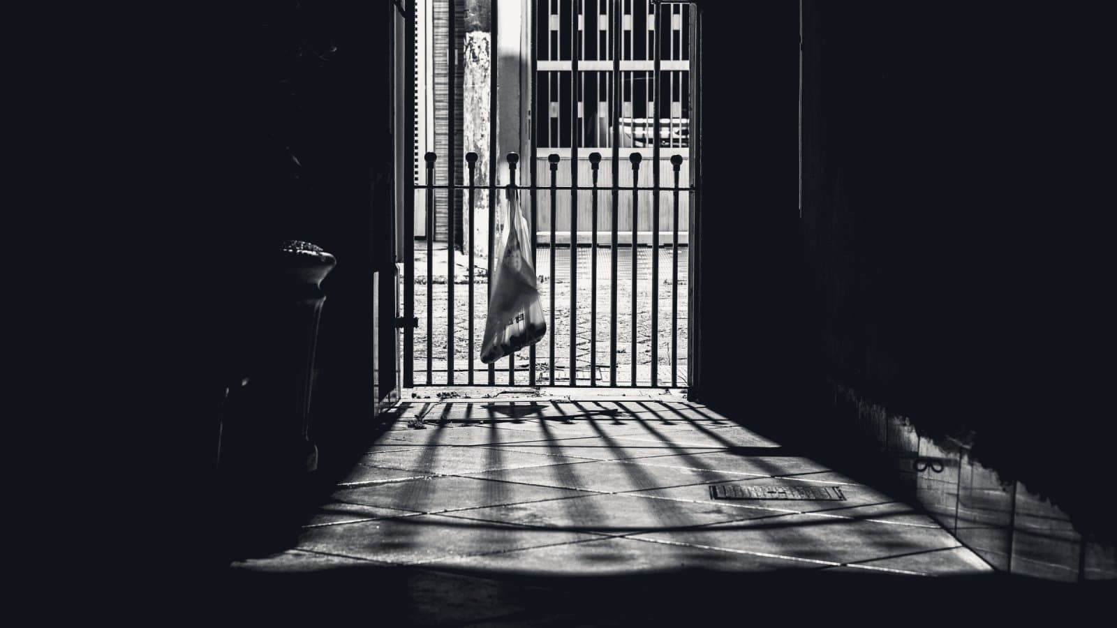 A dark covered passage ending in a barred iron gate, hard daylight beyond it throwing long striped shadows of the bars across a tiled floor.