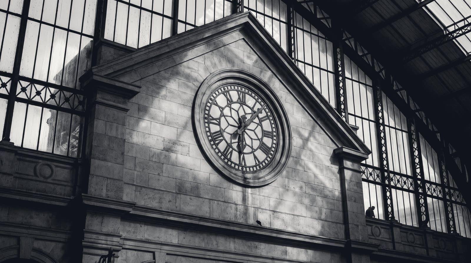 A large exterior clock face set into the stone gable of a railway terminus, read from the concourse below.