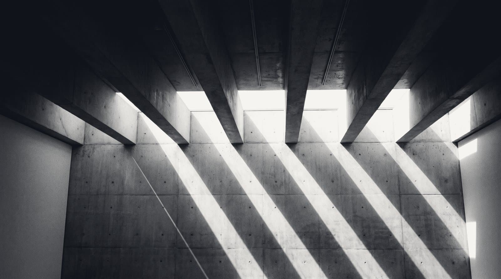 A bare concrete ceiling of deep parallel beams throwing hard stripes of shadow down a gallery wall.