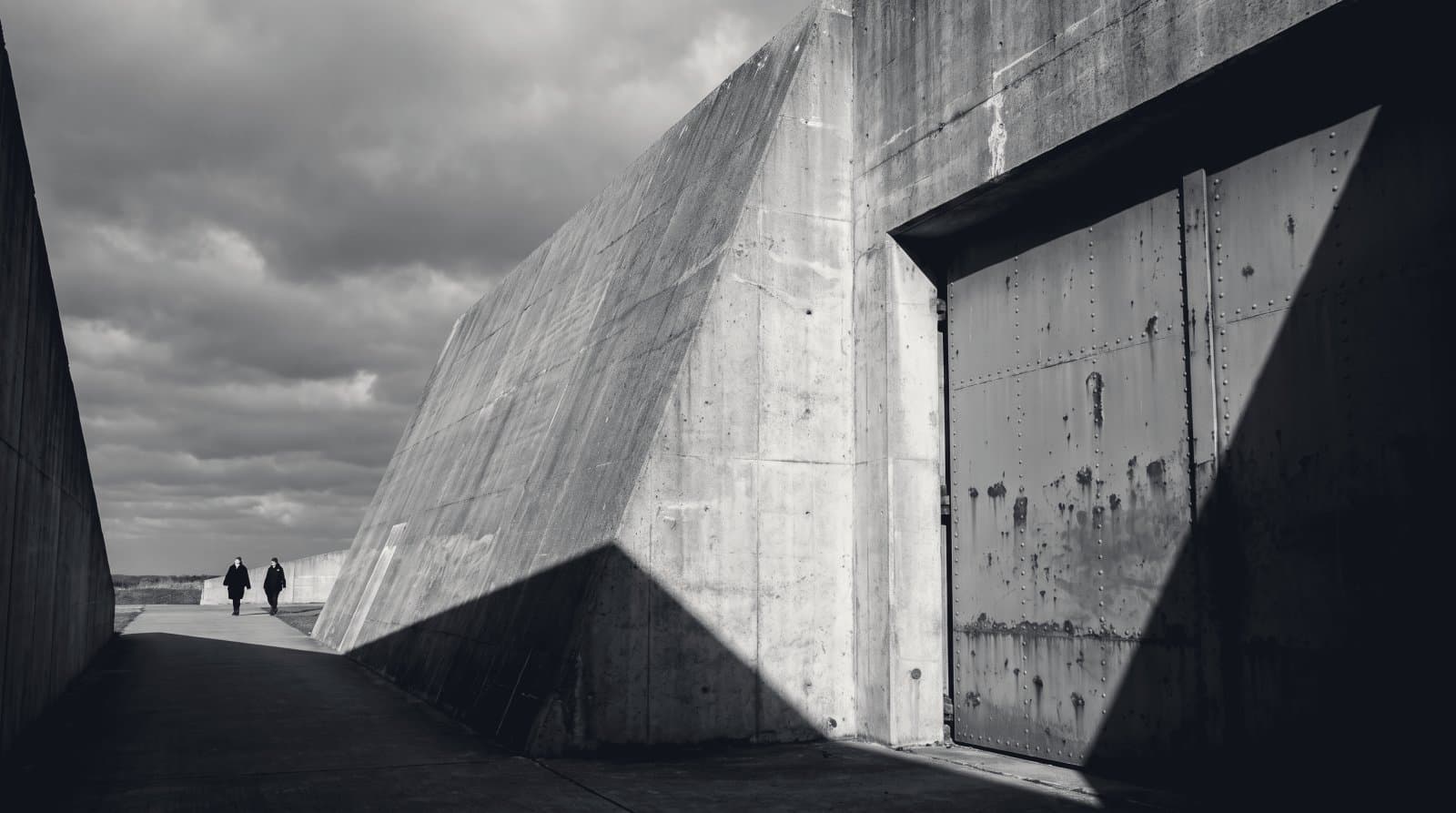 The angled concrete blast wall and steel blast door of a hardened defence facility in hard directional light.