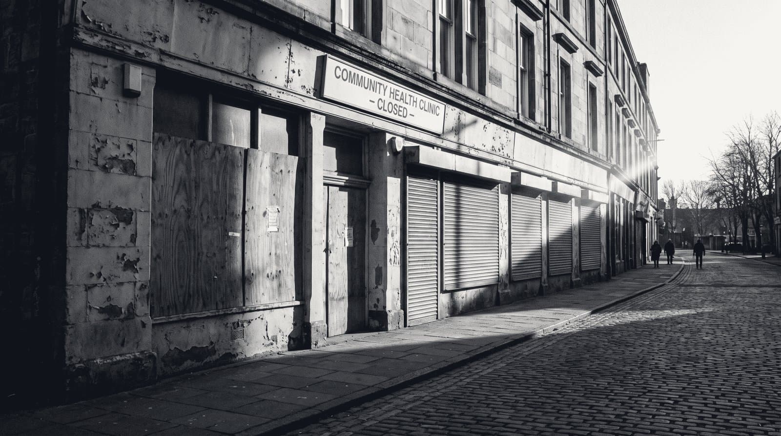 The boarded and shuttered ground floor windows of a closed clinic building on a quiet street.
