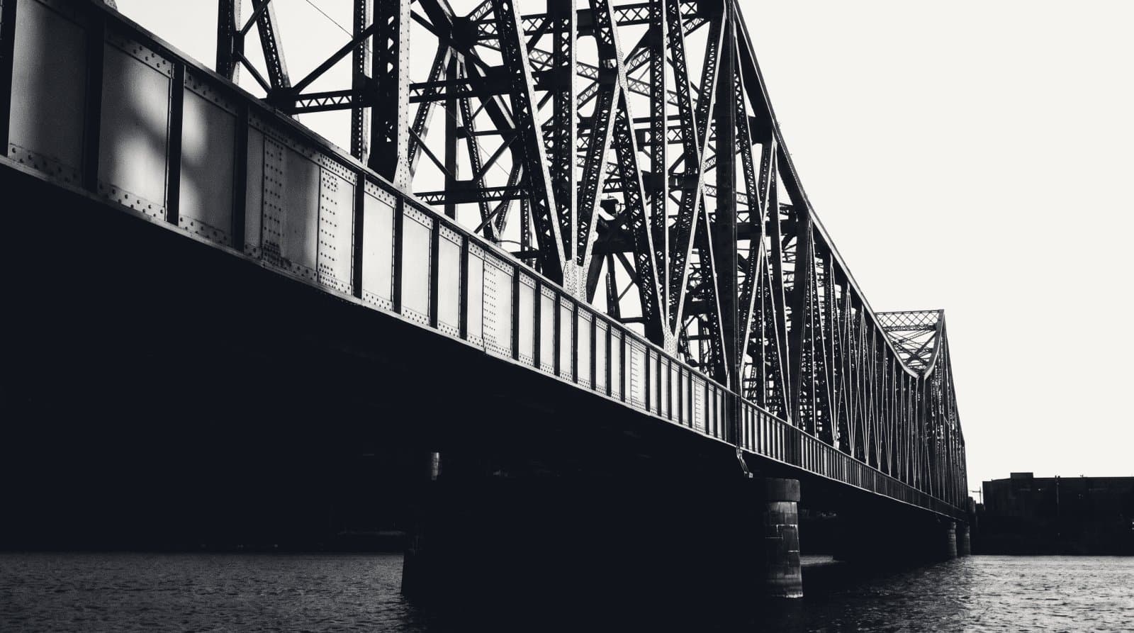 The riveted underside of a steel cantilever bridge span reaching out over water.