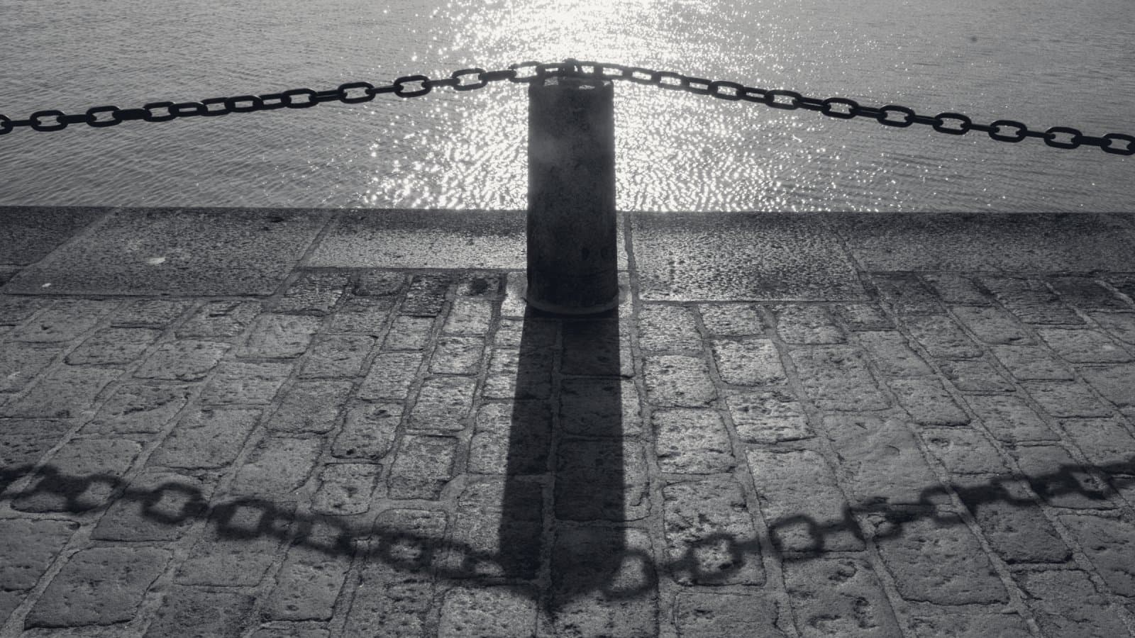 A single dark mooring bollard on a stone quay, backlit by sunlight on the water, with a heavy chain crossing the frame and the chain's shadow drawn long across the cobbles.