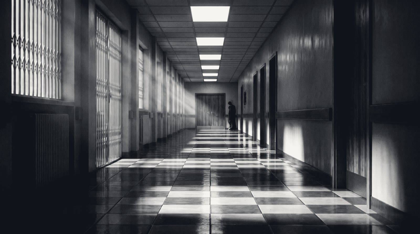 An empty tiled hospital corridor with recessed ceiling lights receding toward a distant closed door.