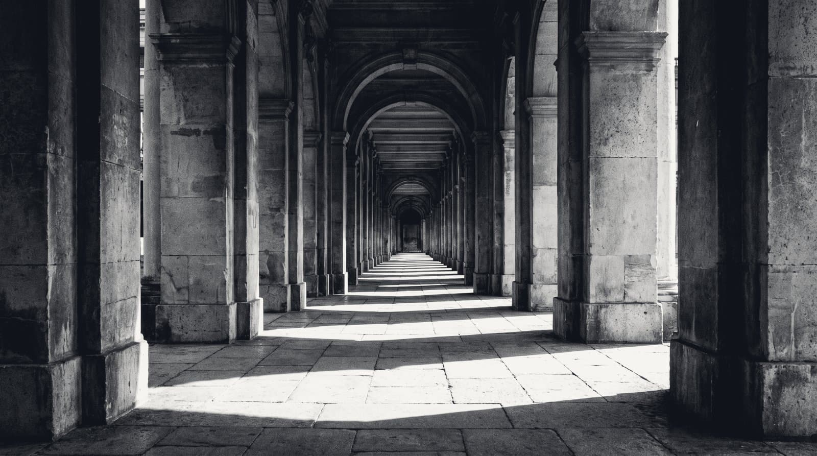 A long stone colonnade receding to a vanishing point, bars of light across the floor.