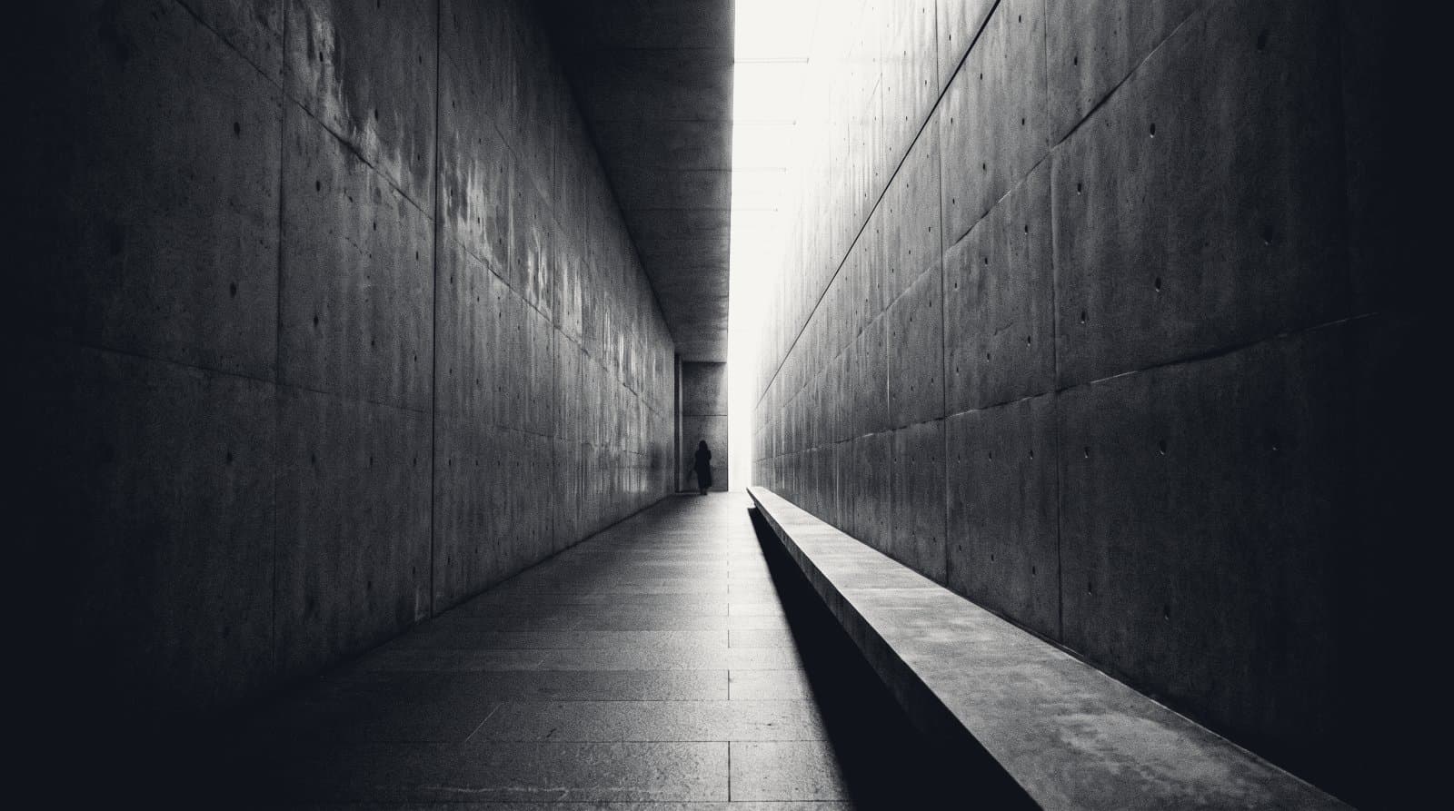 A long passage between two tall concrete walls that lean toward each other, the gap narrowing to a thin bright slot of daylight at the far end.
