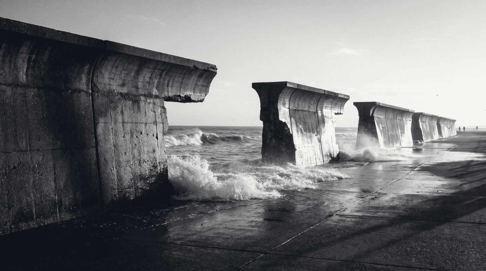 A concrete sea wall with several missing sections, waves running through the gaps onto the promenade.