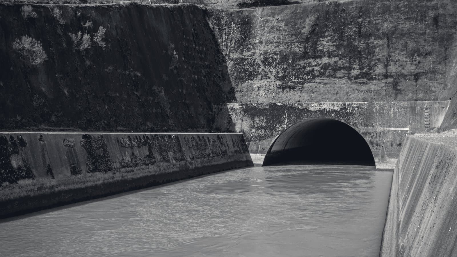 The mouth of a concrete culvert discharging into a straight lined channel between high retaining walls, in hard low sunlight.