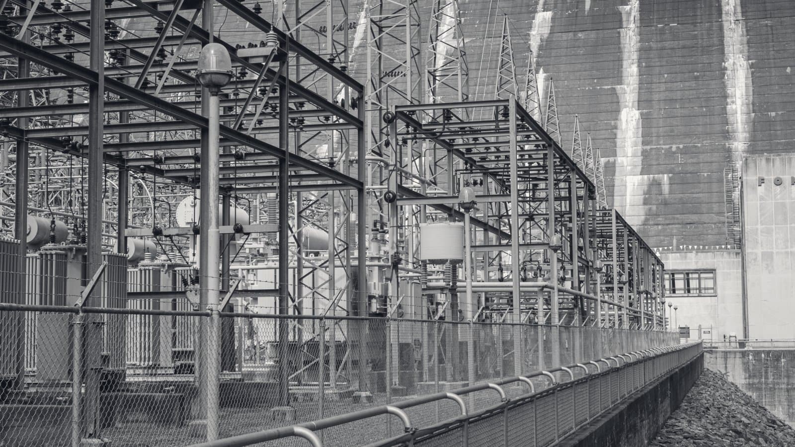 Transformer yard and steel switchgear frames at the foot of a concrete dam, the dam face streaked with water above them, a chain-link fence and railing running along the front.