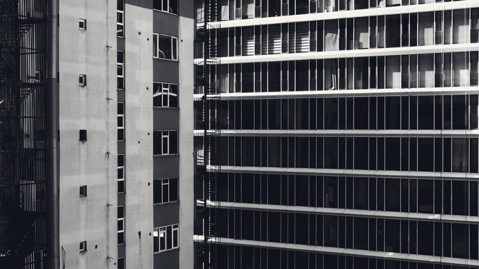 A dense curtain-wall tower with dark, unlit floors beside a plain low block with a fire escape, photographed in hard side light.