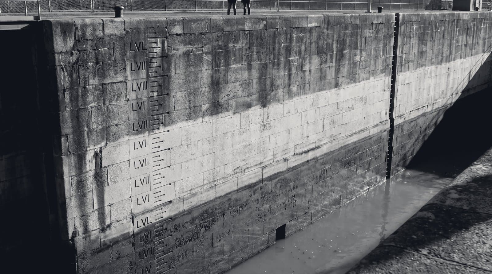 A drained canal lock chamber, its stone wall carved with graduated depth markings, the remaining water sitting far below the upper marks under raking light.