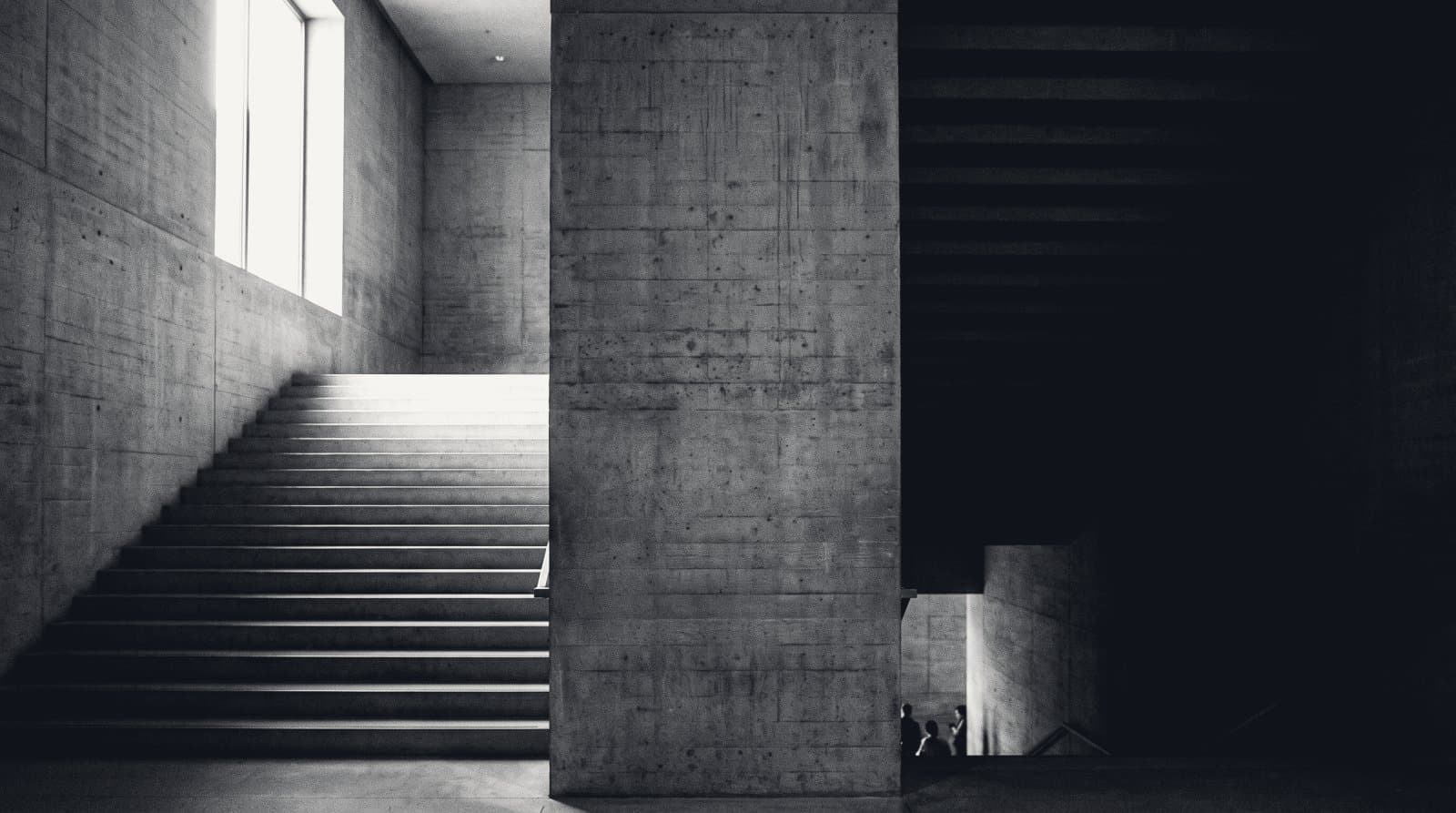 A wide board-formed concrete staircase divided by a massive central pier, the left flight rising into light from a high window and the right flight descending into shadow, with three distant figures at its foot.