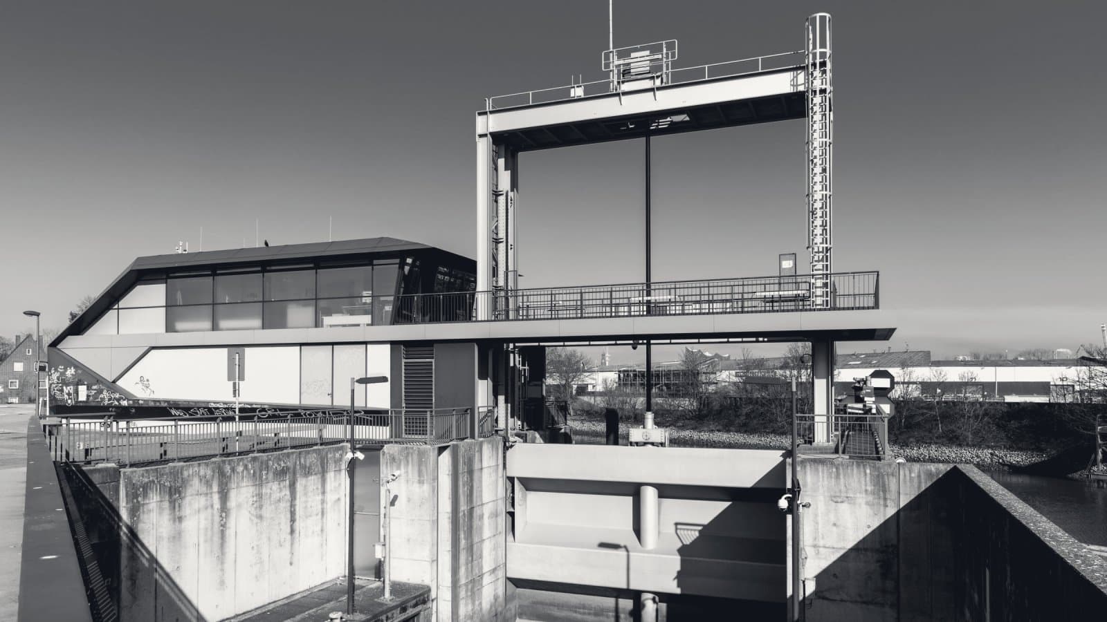 A concrete flood barrier and lock gate under a steel lifting frame, the control house cantilevered above it, in hard low sunlight.