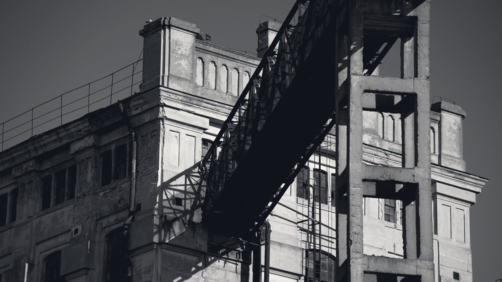 A steel conveyor gantry crossing the upper floors of a weathered stucco industrial building, throwing a hard diagonal shadow down its face.