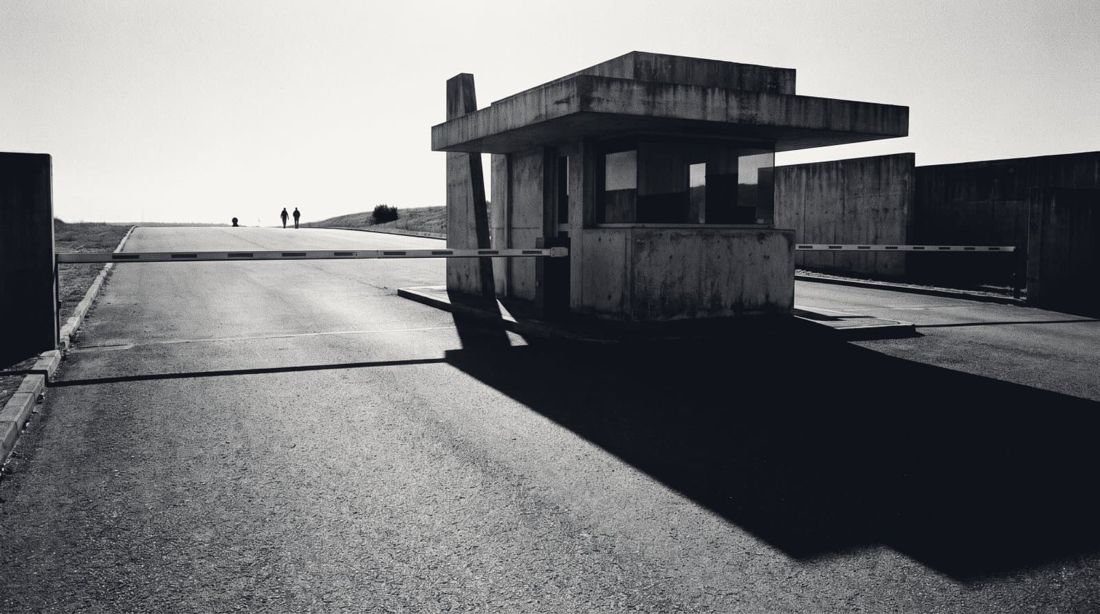 A lowered barrier at a concrete gatehouse controlling a wide empty approach road.