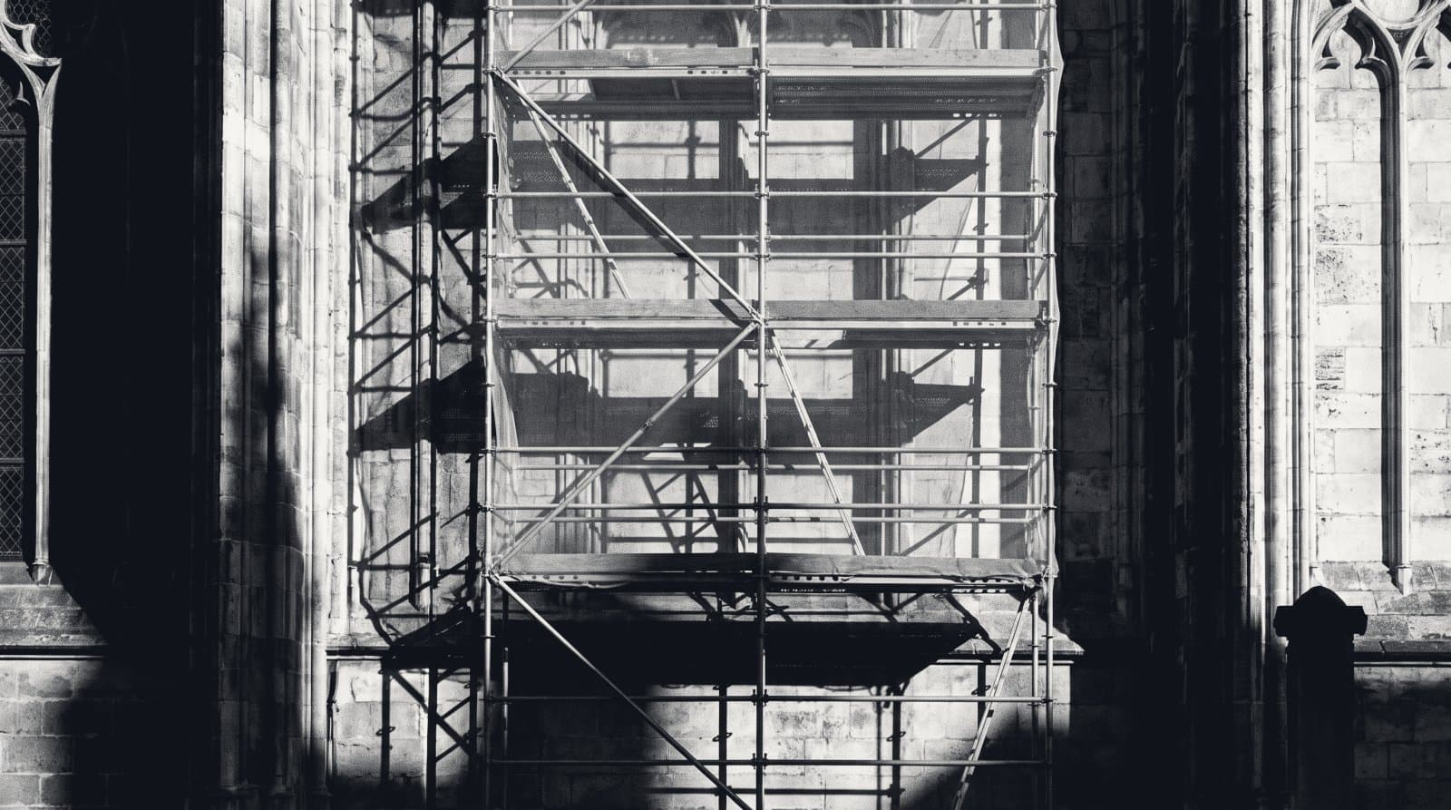 Scaffolding and inspection netting against one bay of a stone facade, casting a lattice of shadow.