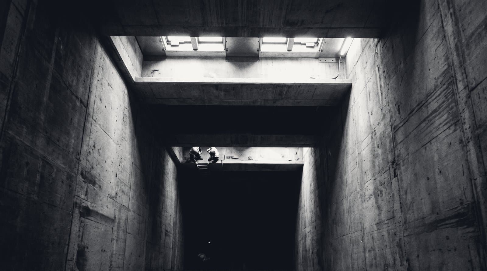Looking along a deep concrete service shaft, a narrow lit ledge crossing it near the top with two distant figures at the edge, and darkness falling away below.