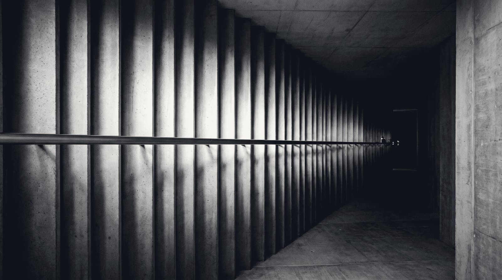 A level steel handrail running dead straight along a fluted concrete corridor while the floor beneath it falls away into shadow.