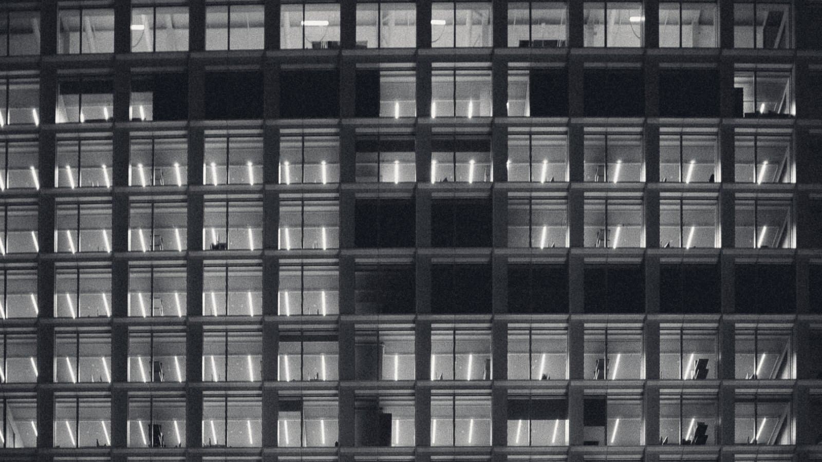 A tall office facade at night filling the frame, hundreds of identical window bays lit from within, a scattered handful of them dark.
