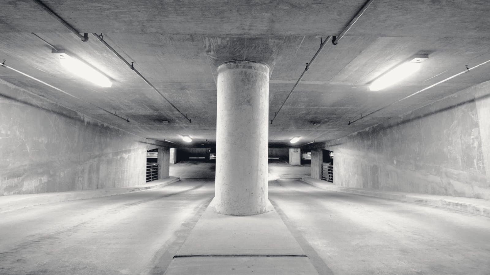 The lowest level of an underground concrete parking structure, a single round column carrying the deck above down the centre of an empty ramp, fluorescent lights receding into shadow.