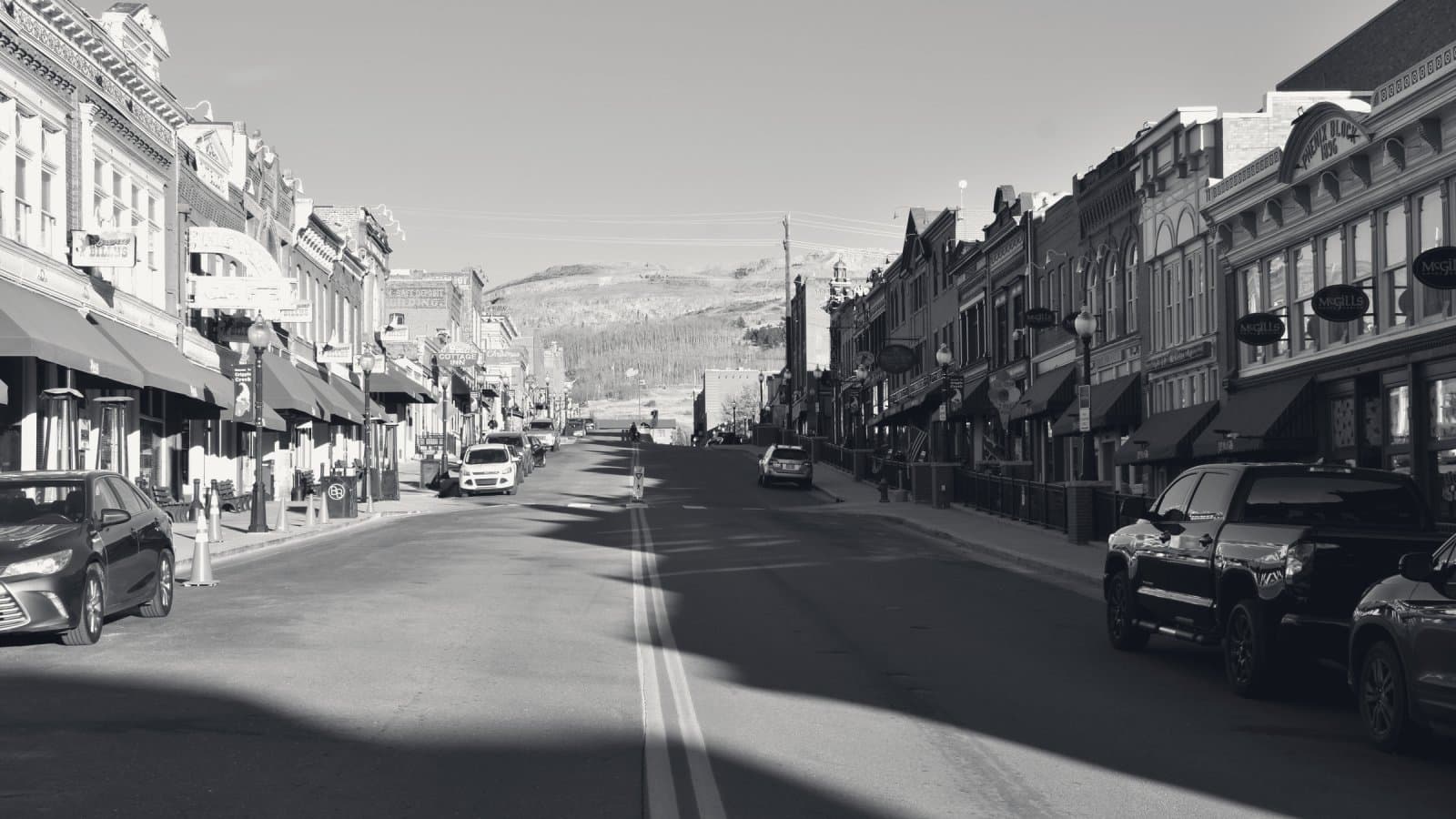 A wide main street of ornate nineteenth-century commercial blocks receding toward a bare hillside, one side in full sun and the other casting a long jagged shadow across the road.