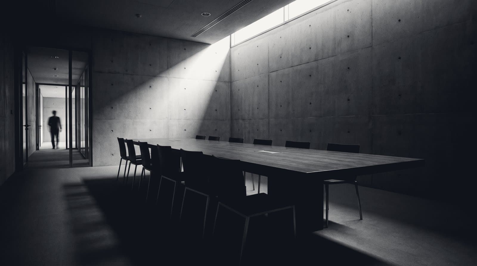 A long empty boardroom table in a concrete room, a shaft of light from a clerestory window, one distant figure in the doorway.