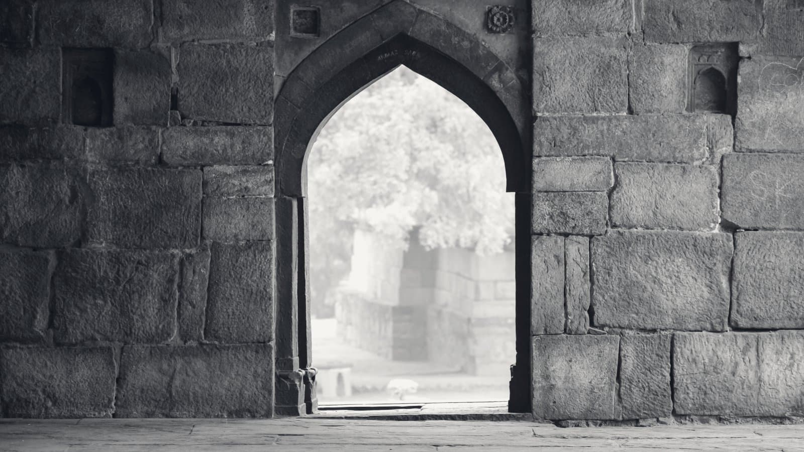A thick wall of large weathered stone blocks pierced by a single pointed archway, bright daylight and a blurred garden beyond it, a worn stone floor in the foreground.