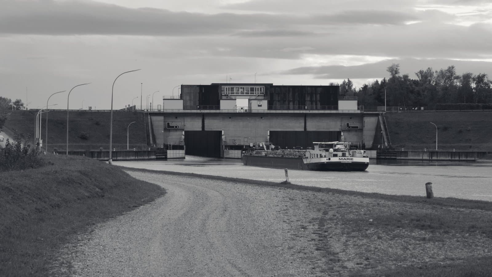 A single long cargo barge on a wide canal approaching a concrete lock with two closed gates, seen from a gravel towpath under a heavy sky.