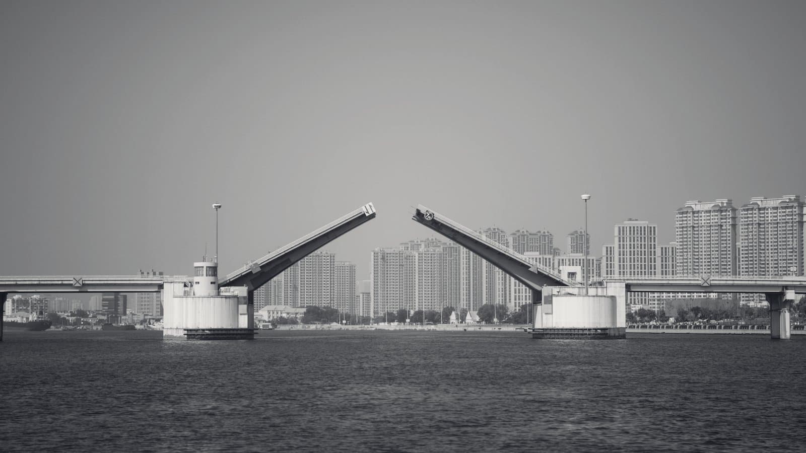 A double-leaf bascule bridge standing open over a wide river, its two raised leaves angled toward each other with a gap of sky between them, apartment towers along the far bank.