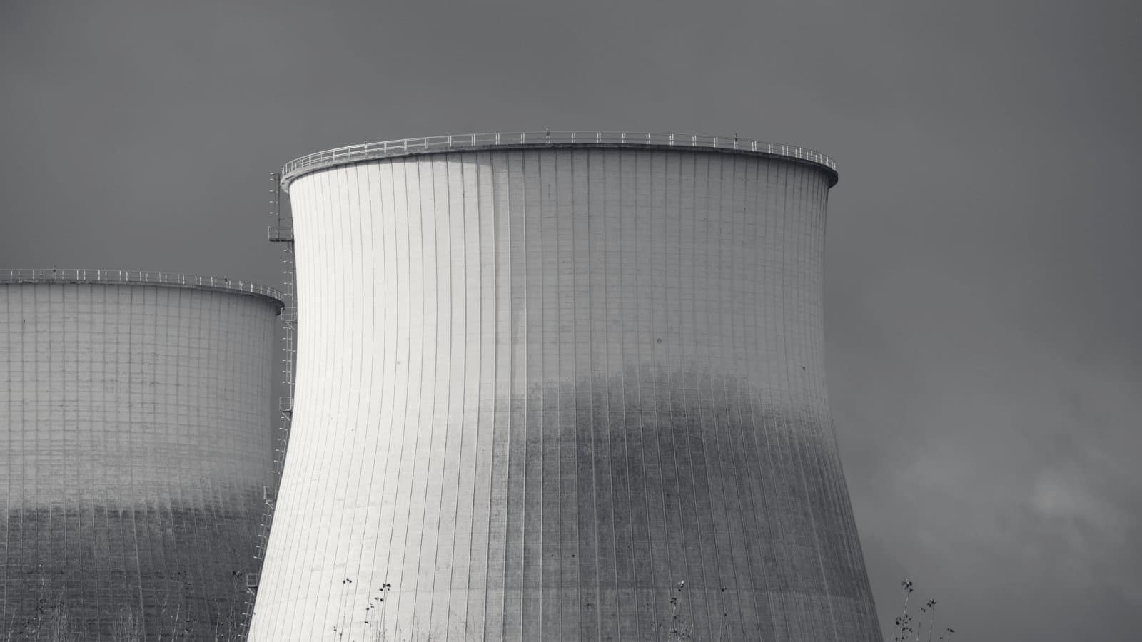 Two very large concrete cooling towers side by side under a dark sky, the nearer one filling most of the frame and lit from one side, thin grasses along the foot of the picture.