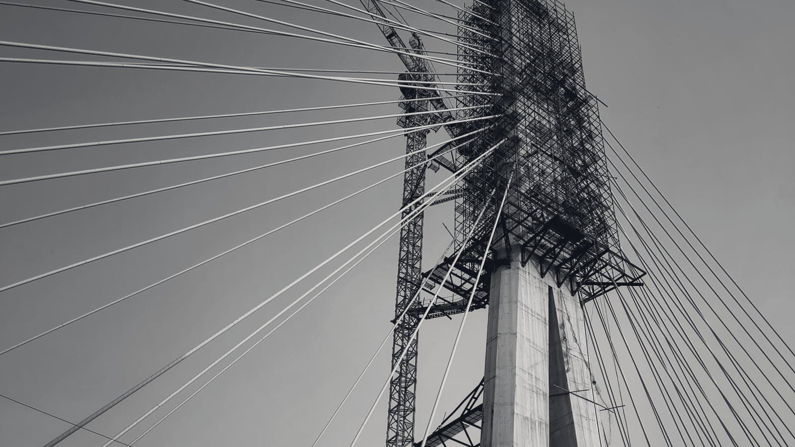 A concrete bridge pylon still wrapped in scaffolding at its head, stay cables fanning out from it and a tower crane rising alongside.