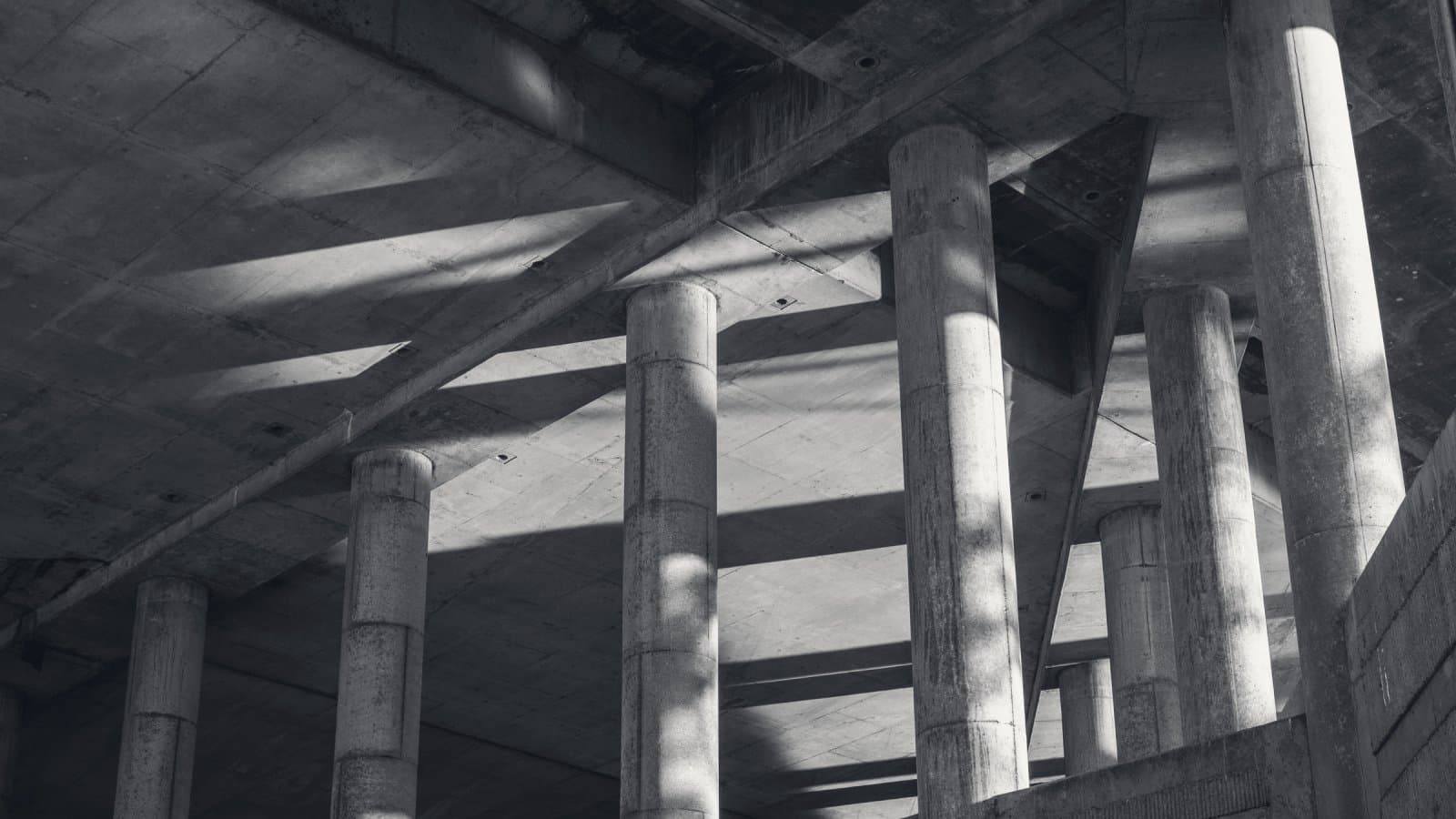 Deep concrete beams under a raked soffit carried on a run of round columns, hard low sunlight cutting across them.