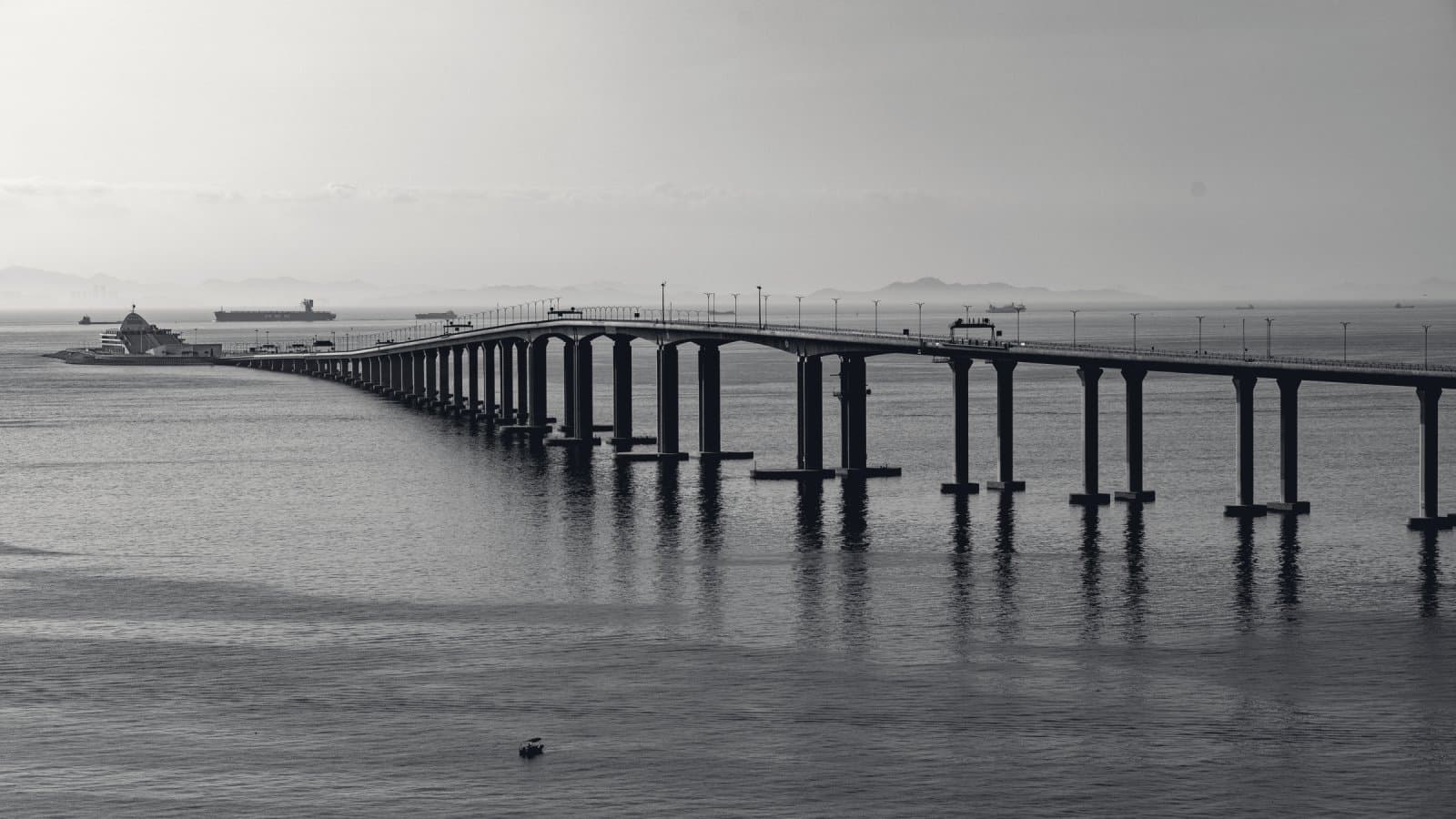 A long, level bridge deck on rows of concrete piers running across open water toward a hazy horizon, with cargo ships in the distance.