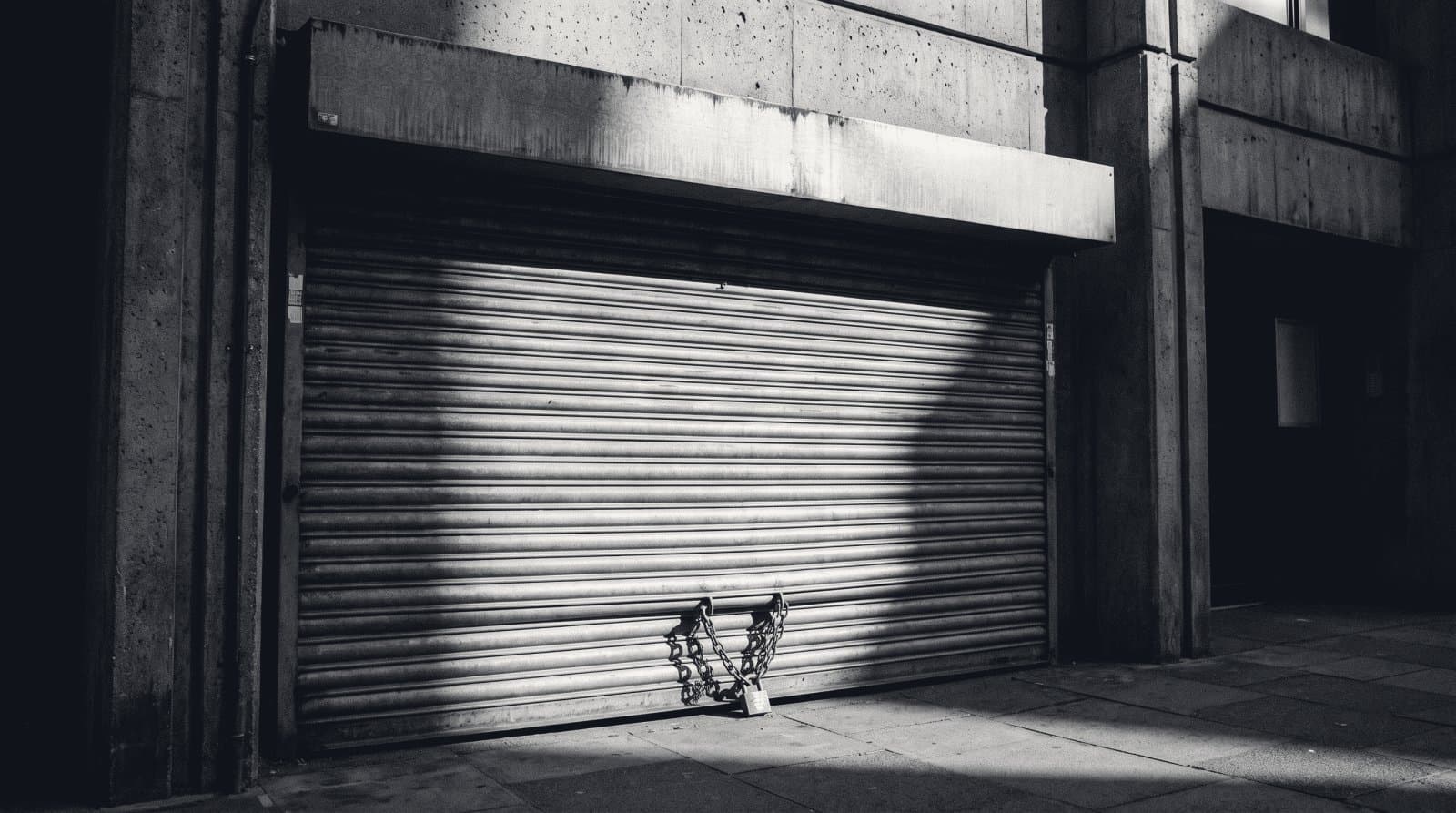 A rolled-steel security shutter closed across a commercial entrance, chain and padlock in hard light.