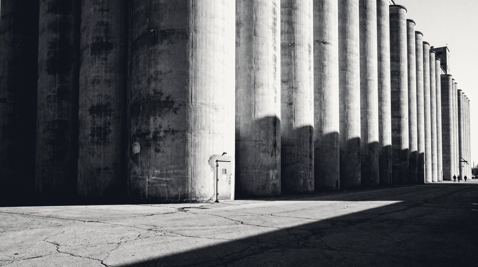 A long receding battery of towering concrete silos in raking light, with one small service door at the base of the nearest cylinder.
