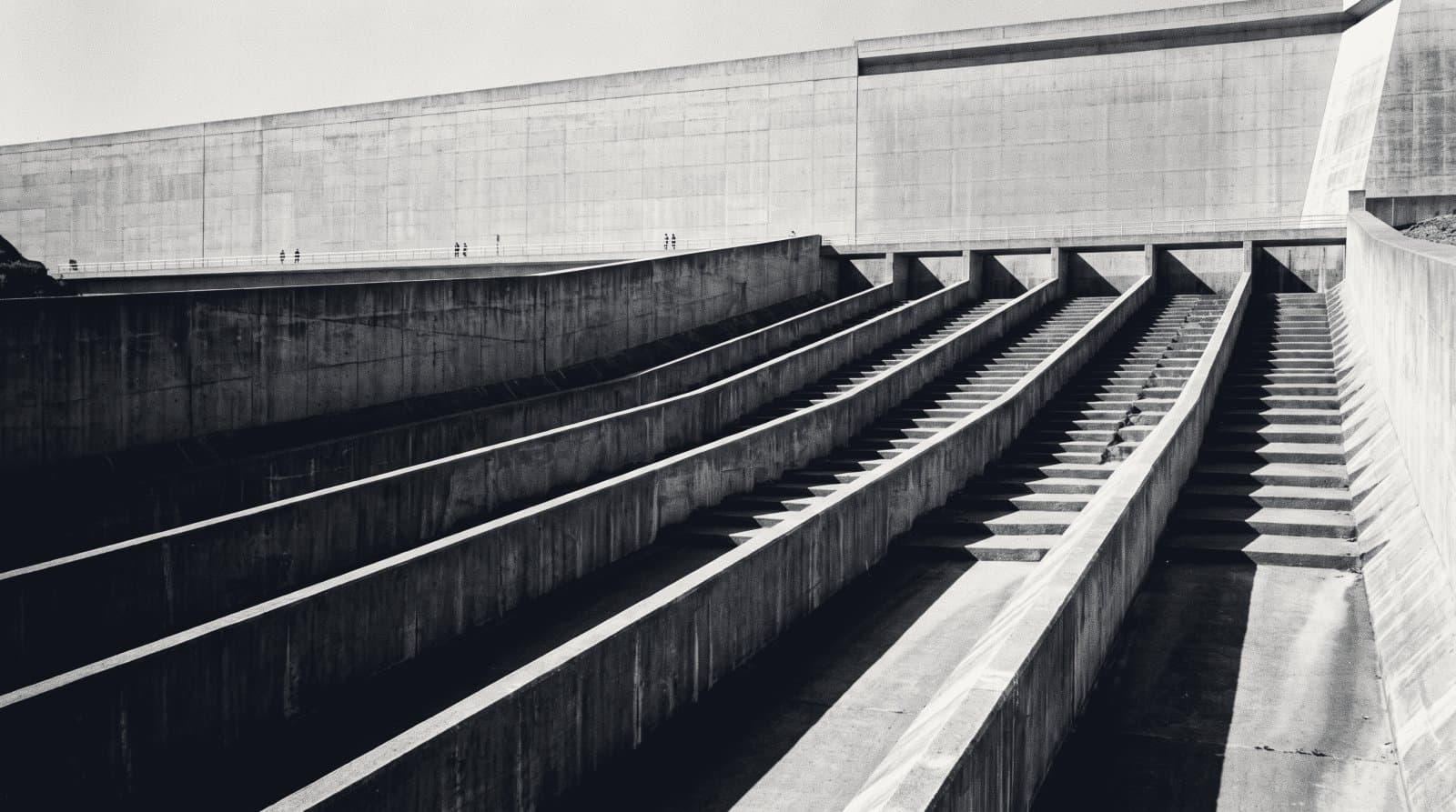 The concrete face of a dam spillway seen from below, wide stepped channels descending dry and empty beneath a long retaining wall.
