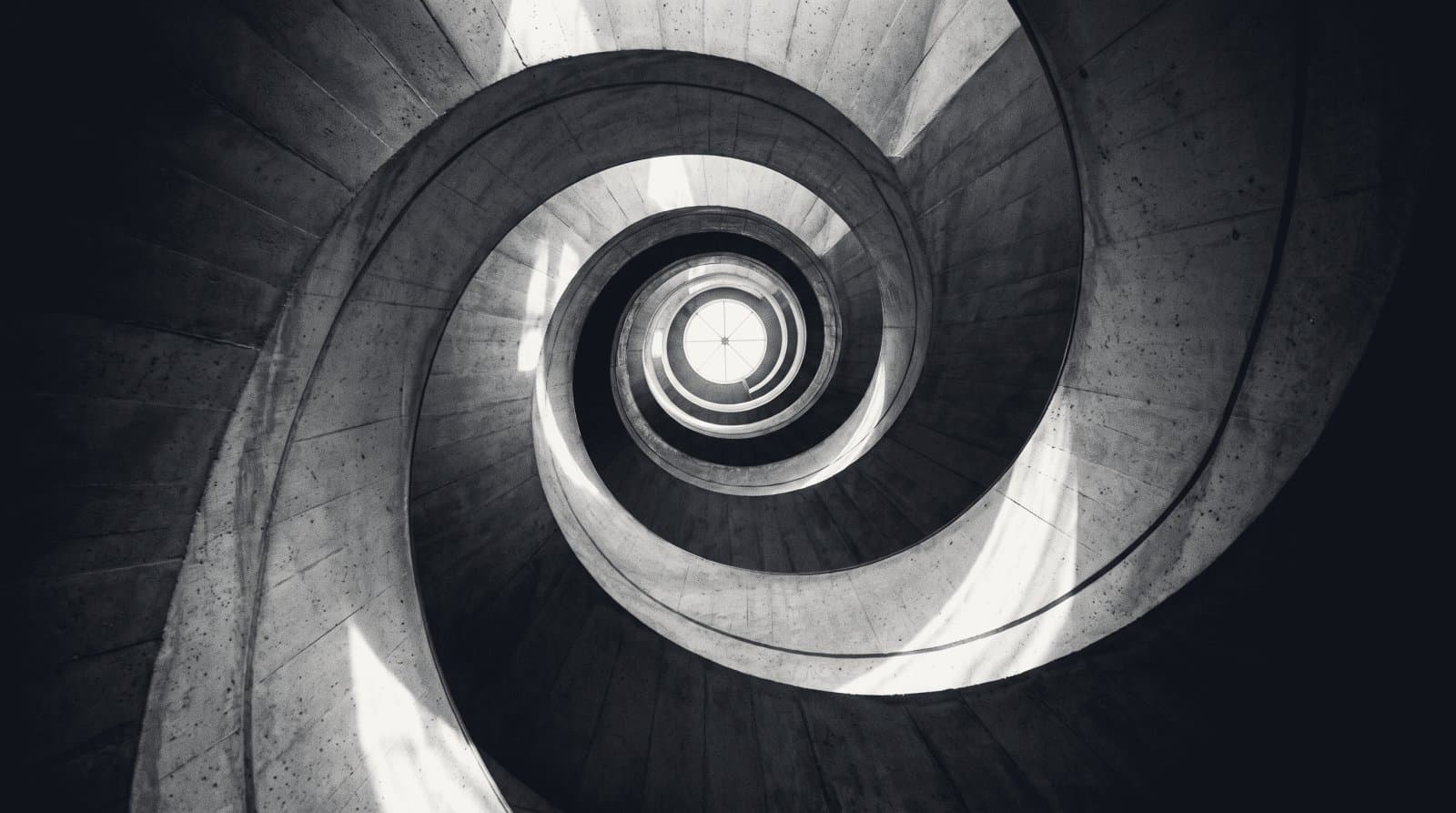 A helical concrete stairwell photographed up the void, turns tightening toward a bright opening.