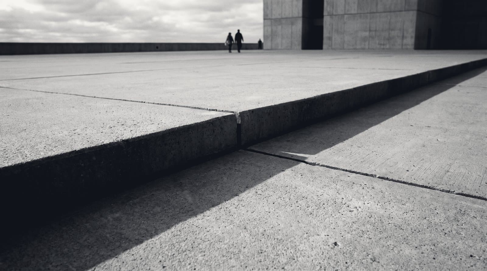 A poured concrete plaza severed by a deep expansion joint, the two slabs settled to different heights, in raking monochrome light.
