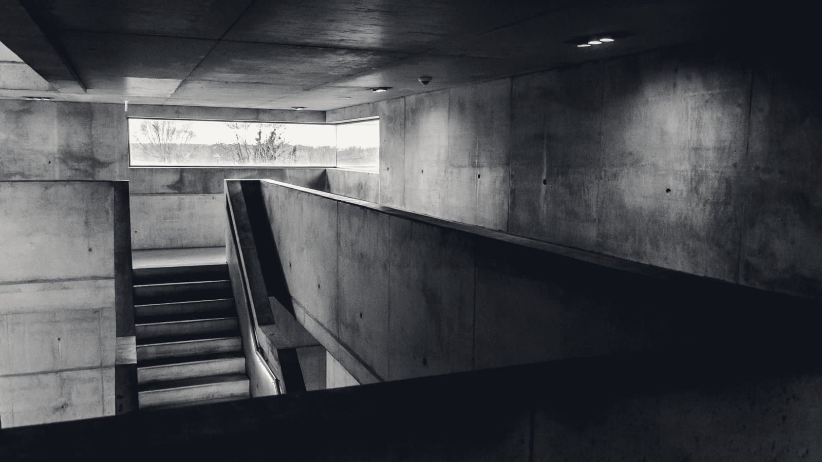 A run of concrete stairs falling away between raw board-marked walls, lit by a single slot window at the turn.