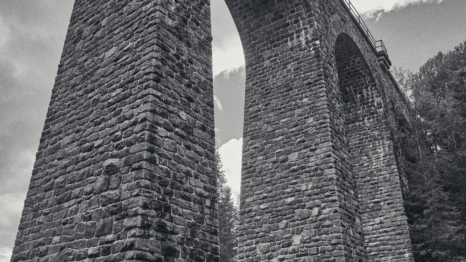 Two stone piers of a railway viaduct seen from below, one arch between them against a clouded sky, forest rising behind the far pier.