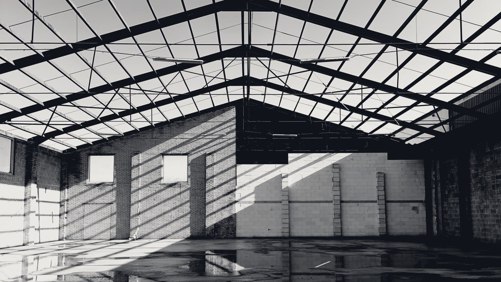 The interior of an empty industrial shed: a steel roof lattice against a bright sky, sunlight striping a brick wall through the gaps, bare block walls, and a wet concrete floor reflecting the beams, with nothing stored inside.