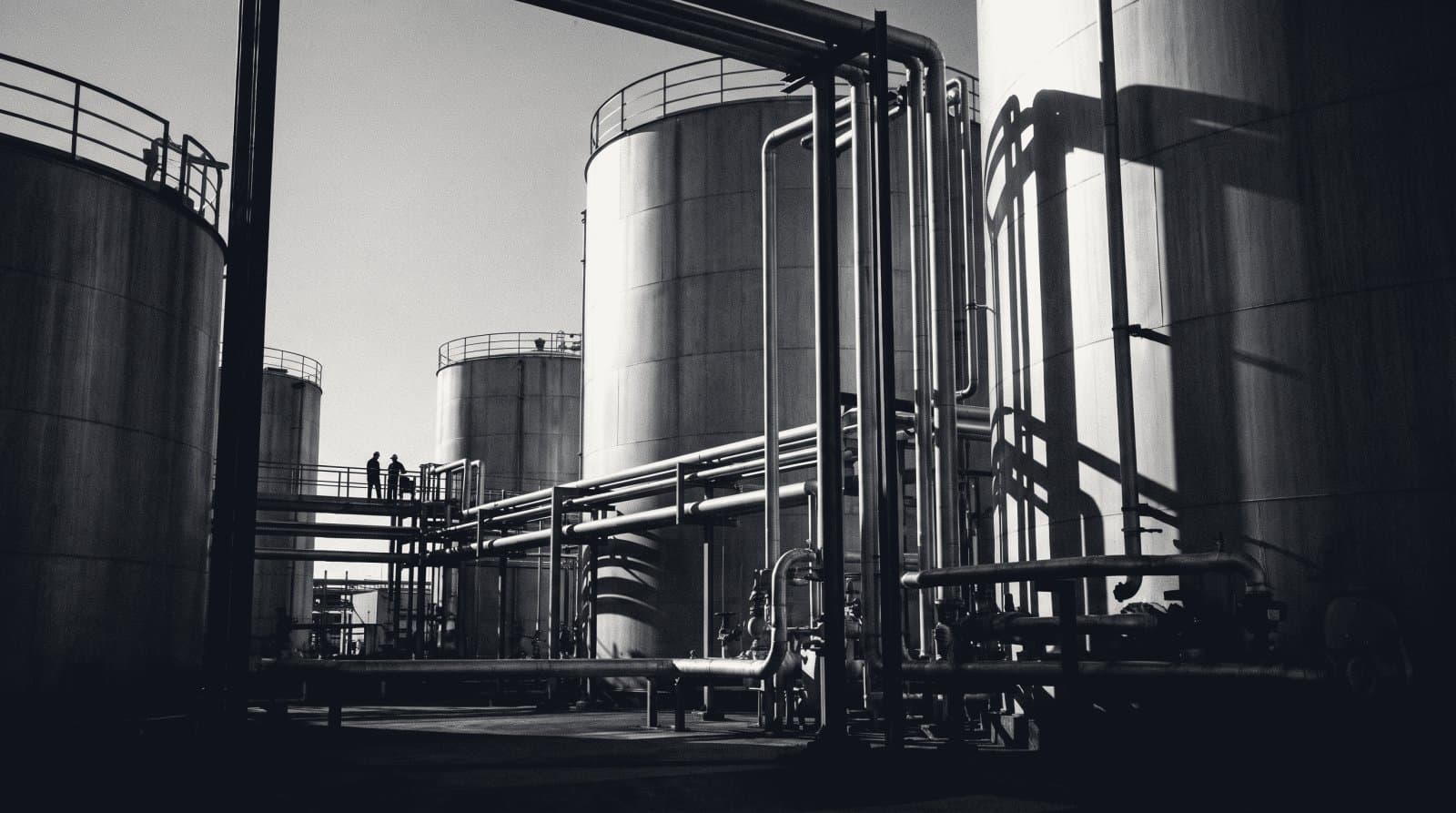 A refinery tank farm of cylindrical storage tanks and pipework seen from a low angle.