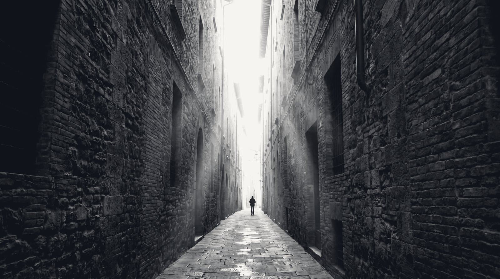 A narrow alley between two tall masonry walls converging toward a bright opening at the far end.