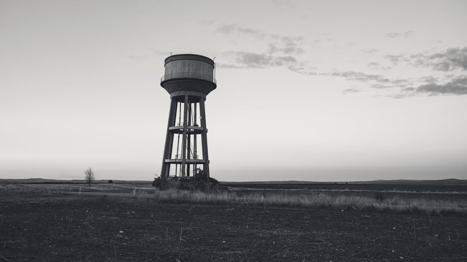 A concrete water tower standing alone on a flat, ploughed plain at dusk, a single bare tree on the horizon to its left.