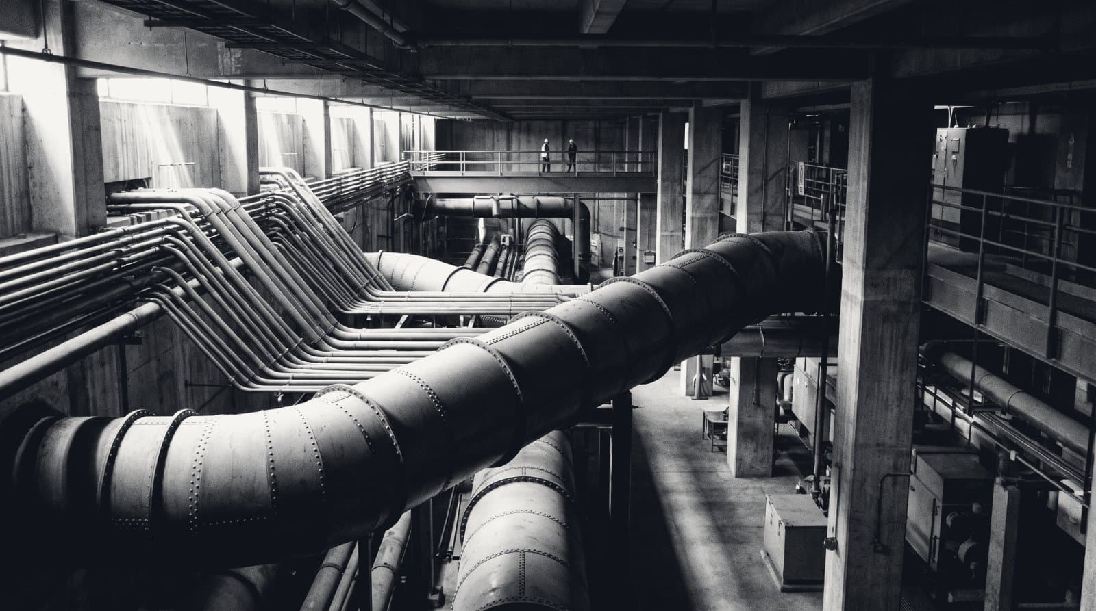 A utility undercroft where dozens of small pipe runs converge into three large riveted trunk mains crossing the frame, raking light on the metal.