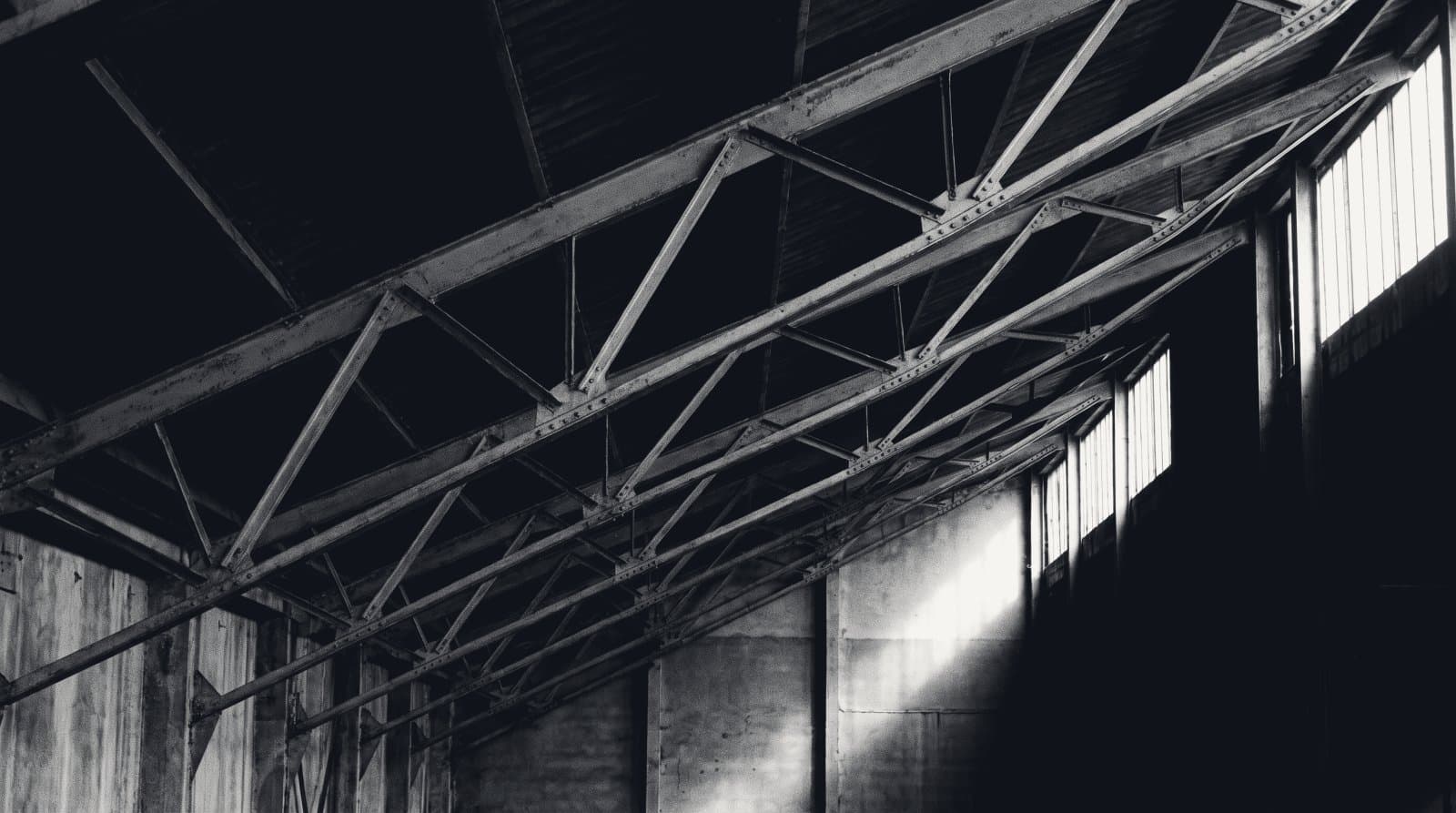 The underside of a deep steel roof truss, repeating diagonal web members receding into shadow.