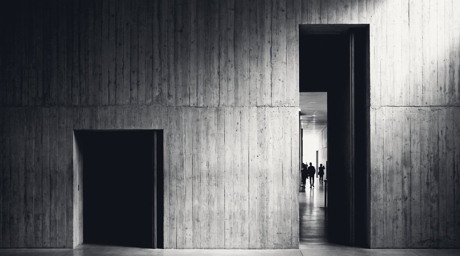 A board-formed concrete wall pierced by two openings, one a tall passage onto a lit corridor with distant figures, the other a lower doorway giving onto darkness.