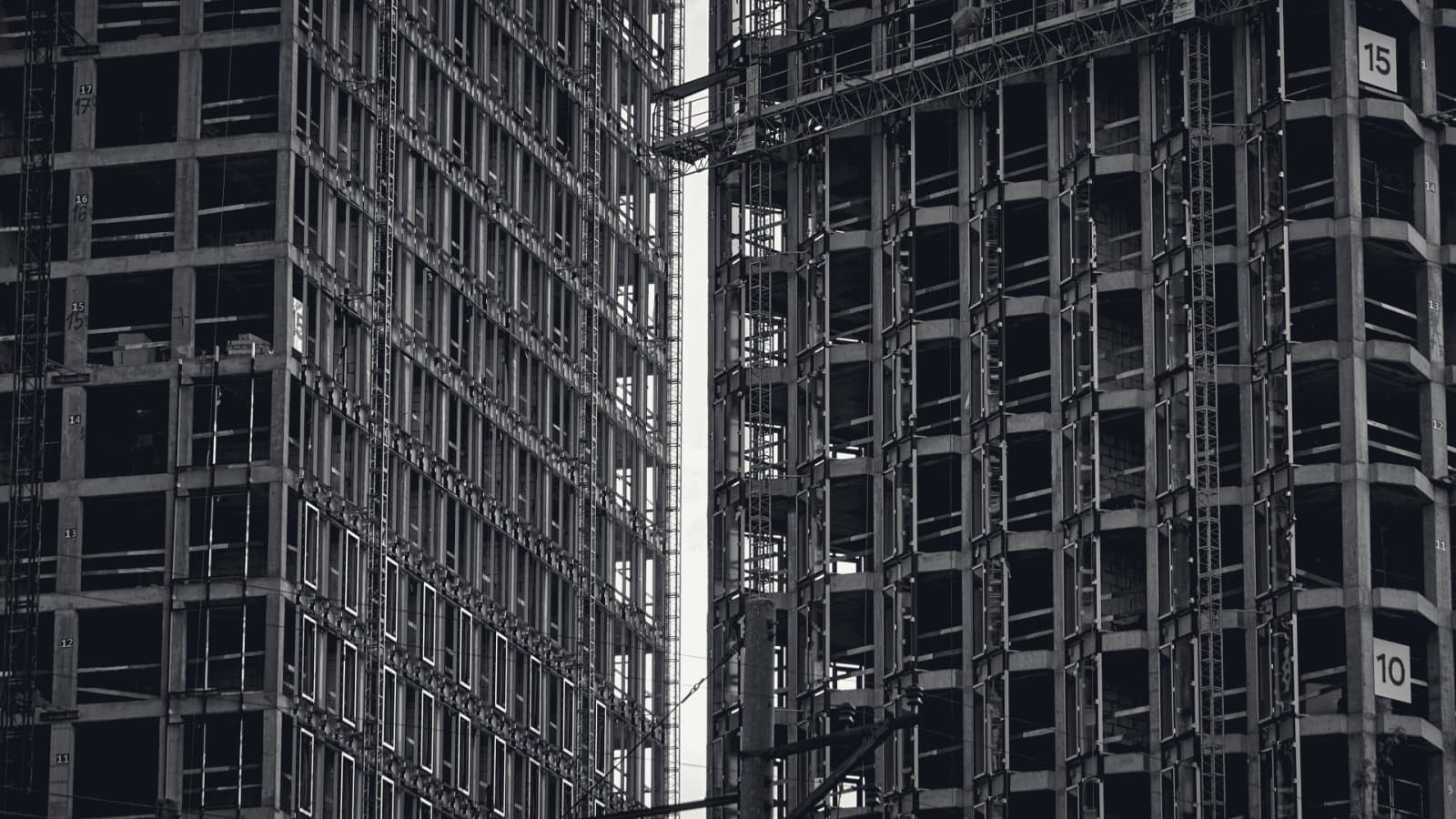 Two unfinished concrete tower frames with open floors and a construction hoist running up the gap between them, floor numbers painted on the columns.